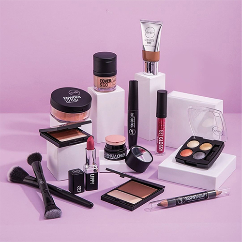 Product shot of cosmetics including powder, lipstick, lip gloss, mascara, brow color, brushes, and foundation