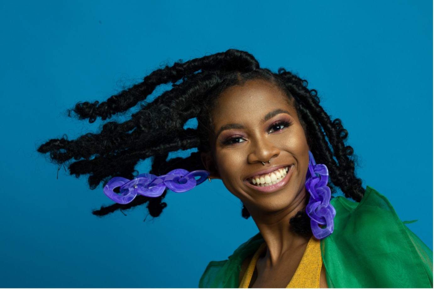 portrait of a smiling Black woman with natural textured hair in twists, bold makeup, large purple chain earrings, green outfit, blue background
