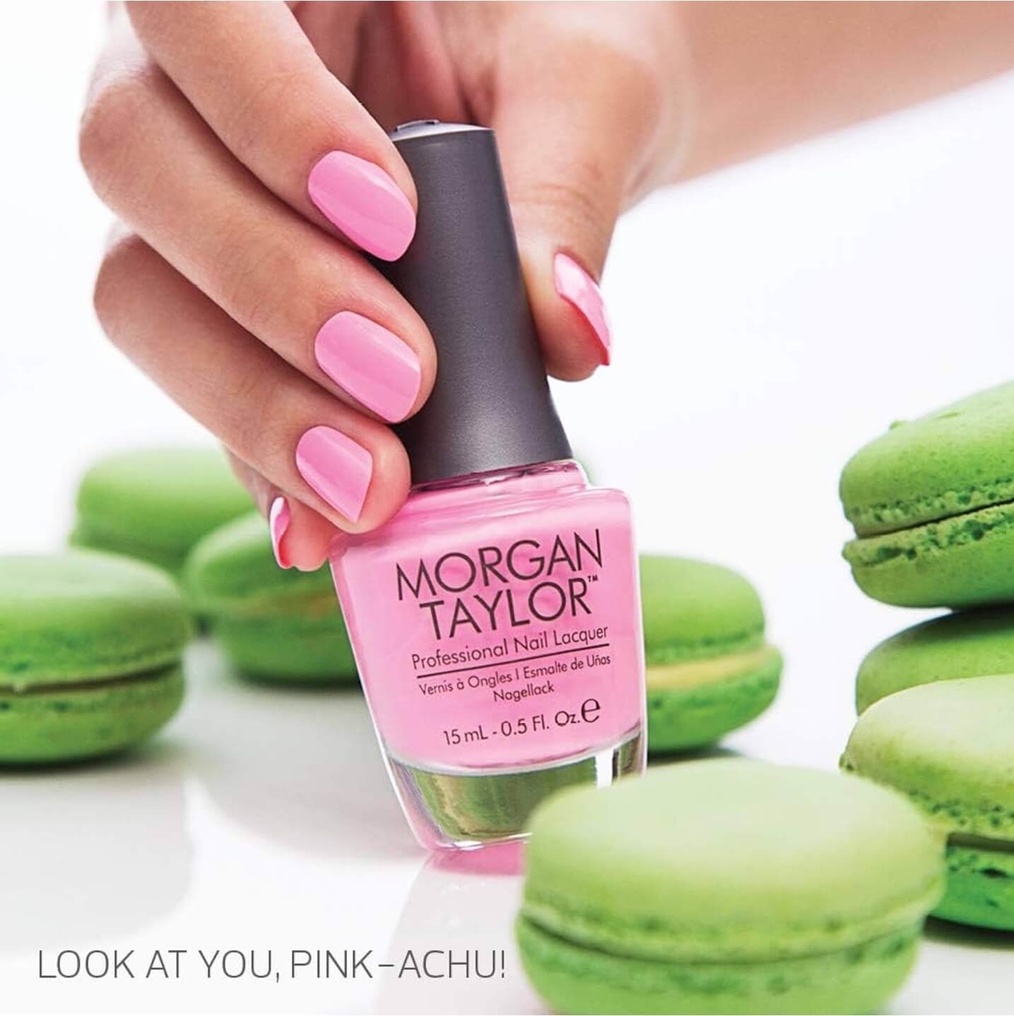 Hand with a pink manicure holding a bottle of Morgan Taylor Professional Nail Lacquer, with green macarons in the background