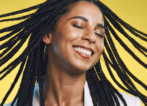 Smiling woman with long braids and hoop earrings against a bright yellow background.